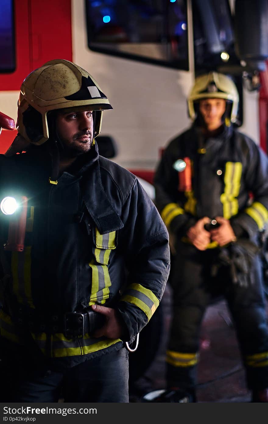 Two brave firemen wearing a protective uniform standing next to a fire truck. Arrival on call at night time