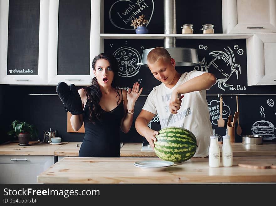 Happy young couple of people standing at kitchen and eating fruits. Beautiful brunette pregnant women holding plate and standing near husband. Handsome men cutting watermelon and sharing it with wife. Happy young couple of people standing at kitchen and eating fruits. Beautiful brunette pregnant women holding plate and standing near husband. Handsome men cutting watermelon and sharing it with wife.