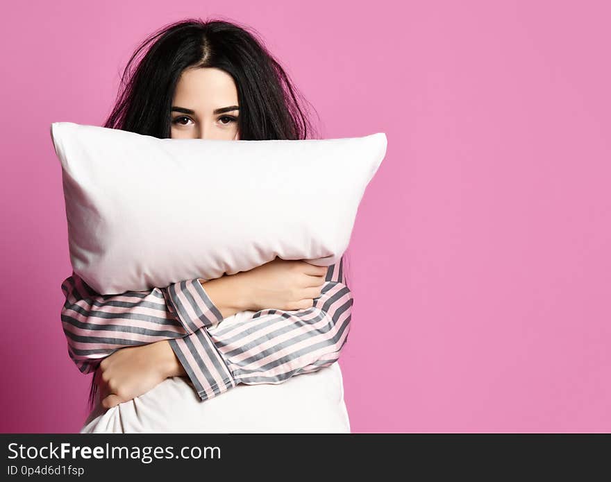 Beautiful brunette girl just woke up with soft pillow feathers and sleepy mask bandage for sleep on pink background