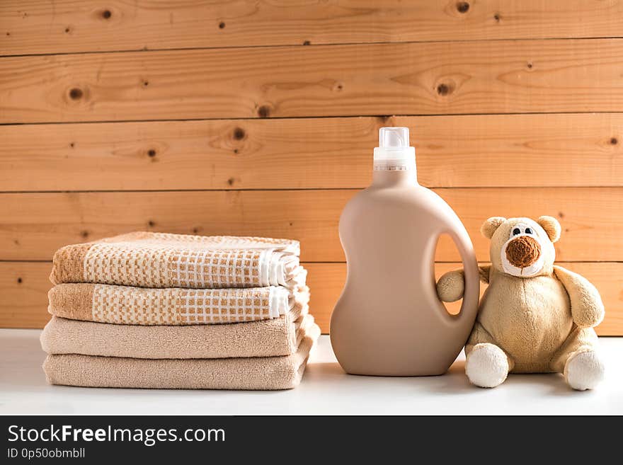 Gel for laundry washing in plastic bottle near a pile of fresh towels. Teddy bear holds detergent bottle. Composition on white wooden shelf against wooden background. Gel for laundry washing in plastic bottle near a pile of fresh towels. Teddy bear holds detergent bottle. Composition on white wooden shelf against wooden background.