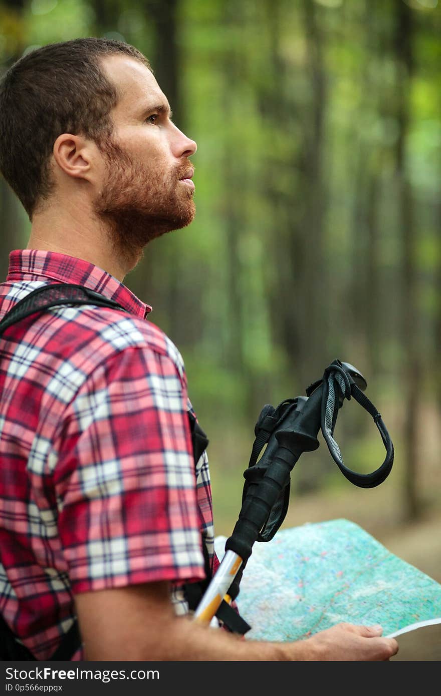 Determined young man in red checkered shirt hiking through lush green forest, holding a map and looking into distance. Determined young man in red checkered shirt hiking through lush green forest, holding a map and looking into distance