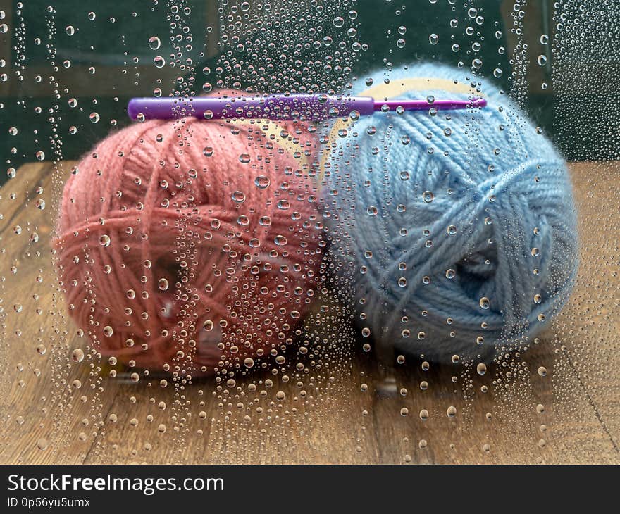 Blue and pink skeins of wool with a purple crochet hook. Seen through a window with raindrops