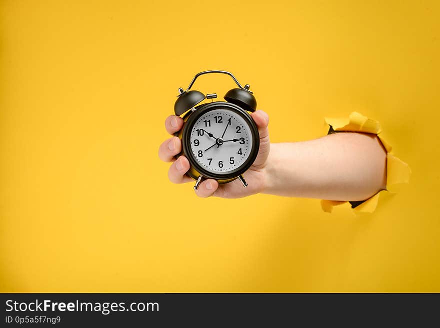 Hand holding a vintage black alarm clock through torn yellow paper background. Wake up on time. Hand holding a vintage black alarm clock through torn yellow paper background. Wake up on time.