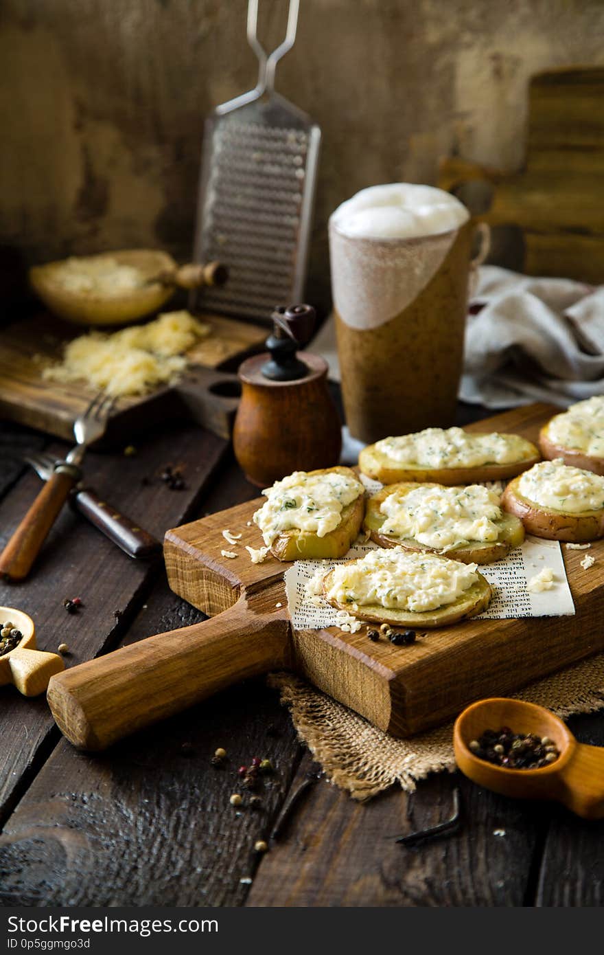 Homemade baked slices of potatoes with cheese with dill, pepper on wooden board standing on rustic table with sackcloth, jar with foam beer, pepper mill, plates with pepper and cheese, forks, towel