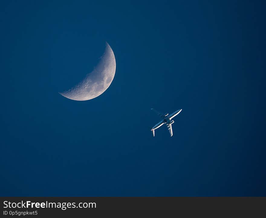 A low-flying commercial airliner passes by a crescent moon in a dark blue sky at sunset. A low-flying commercial airliner passes by a crescent moon in a dark blue sky at sunset