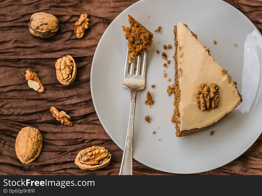 Top view of homemade carrot cake with cream cheese topping decorated with piece of walnut placed on white plate with silver fork on brown tablecloth with walnuts and nutshell next to the plate