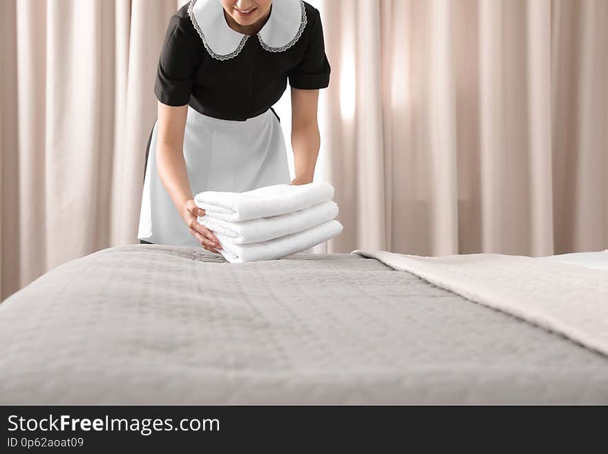 Young maid putting stack of fresh towels on bed in hotel room