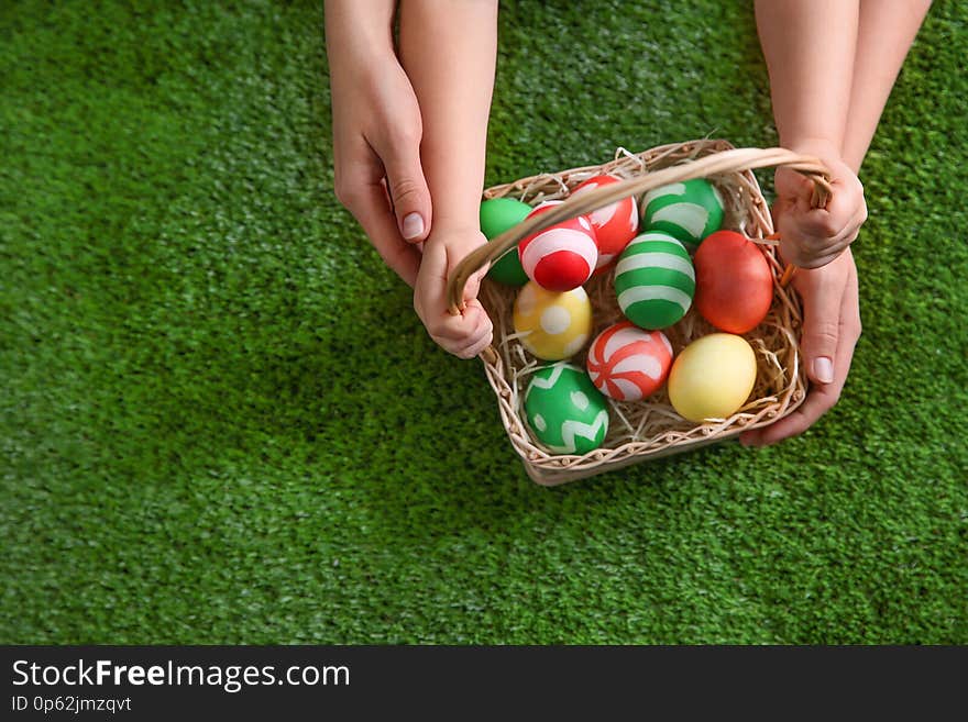 Mother and her little child with basket of painted Easter eggs on green grass, top view. Space for text