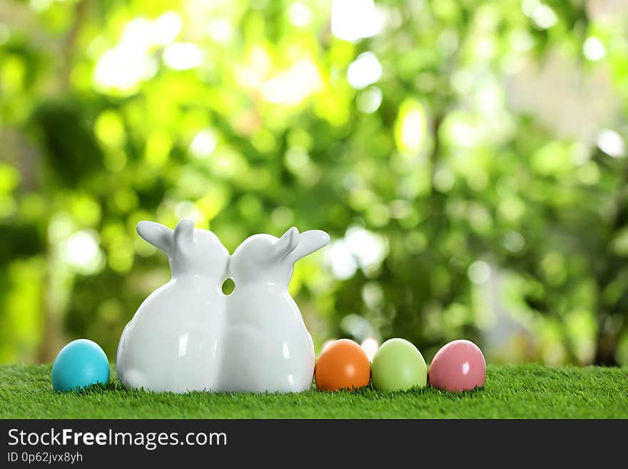 Cute ceramic Easter bunnies and dyed eggs on grass against blurred background, space for text