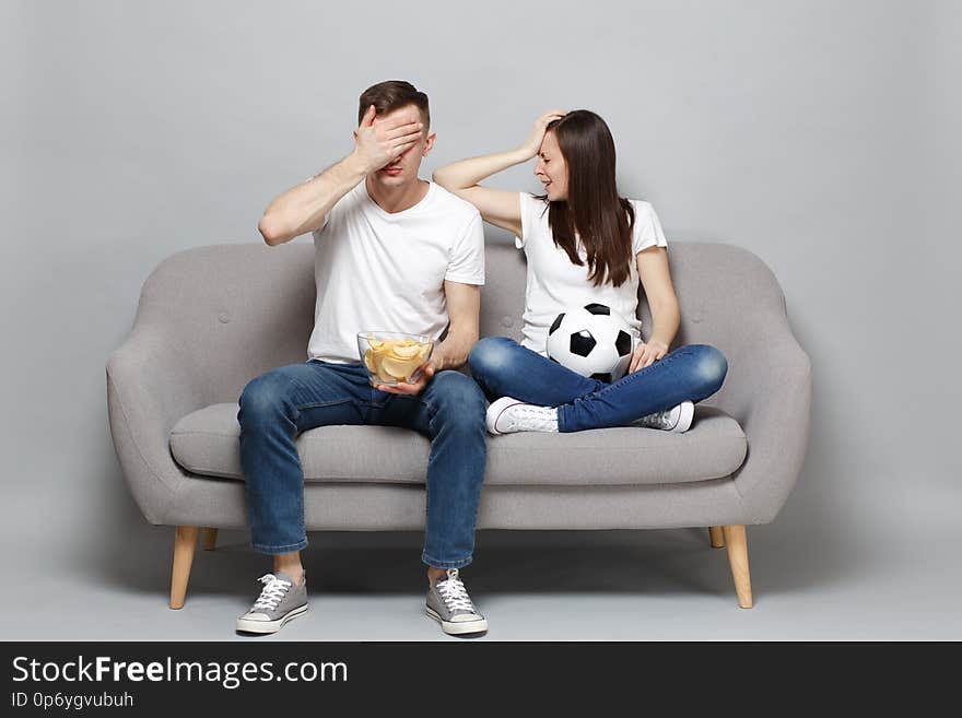 Displeased couple women men football fans cheer up support favorite team with soccer ball, holding bowl of chips, covering face with palm isolated on grey background. Sport family lifestyle concept. Displeased couple women men football fans cheer up support favorite team with soccer ball, holding bowl of chips, covering face with palm isolated on grey background. Sport family lifestyle concept