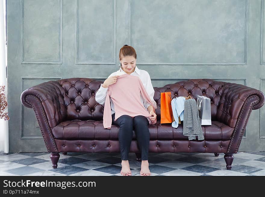 Beautiful Asian woman sitting on the couch.The pink shirt is in his hand.Many shirts And several paper bags on the brown sofa in the room.Woman smiling.Do not focus on the object. Beautiful Asian woman sitting on the couch.The pink shirt is in his hand.Many shirts And several paper bags on the brown sofa in the room.Woman smiling.Do not focus on the object