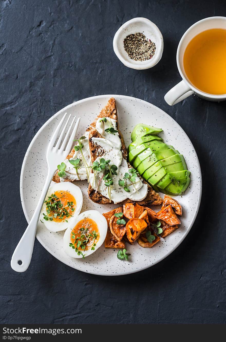 Cream cheese toast, avocado, boiled egg, baked sweet potatoes and tea - delicious healthy breakfast or snack on a dark background, top view