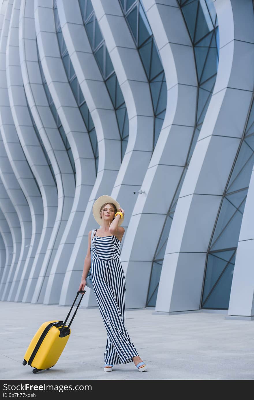 Young stylish Caucasian woman at the airport with a suitcase and a straw hat.