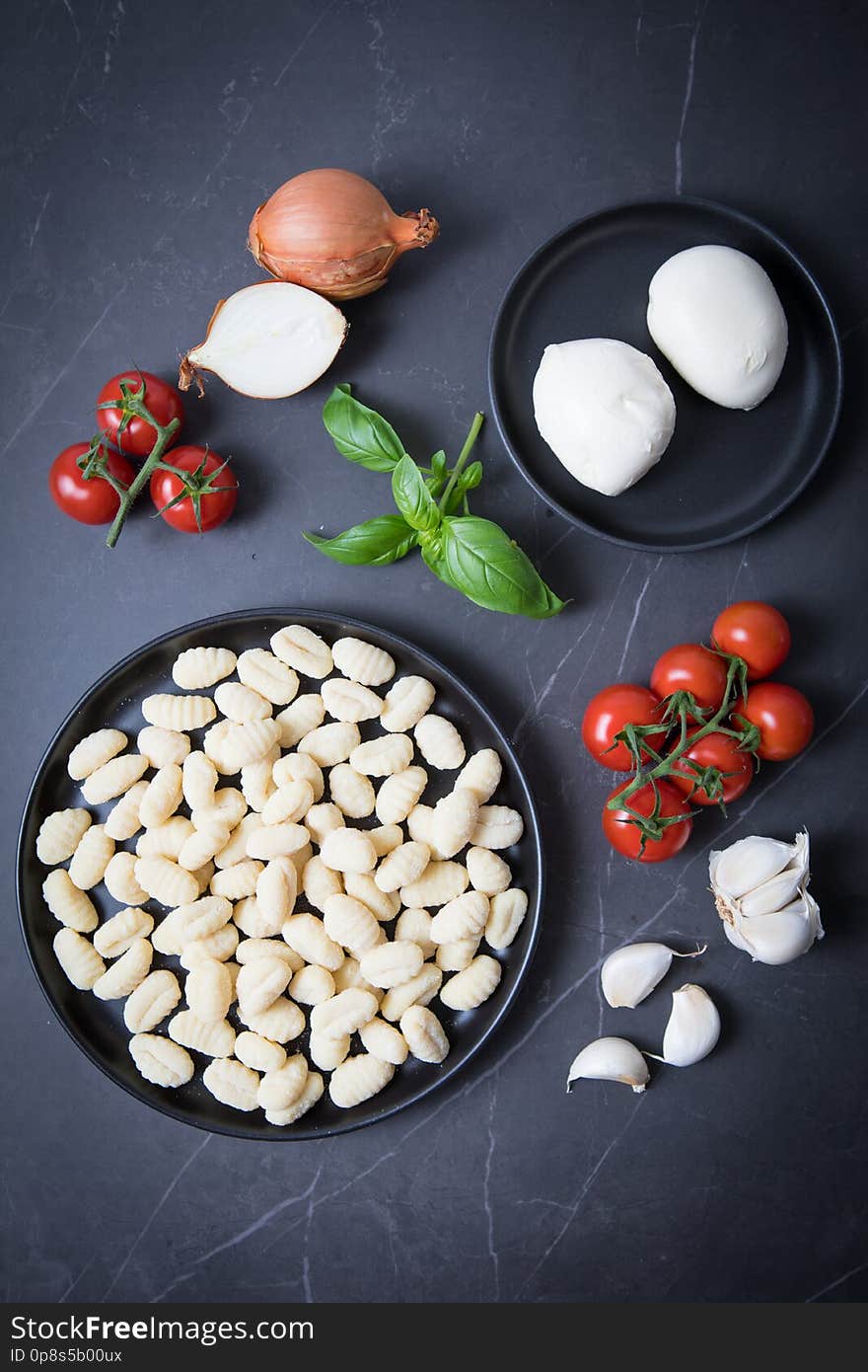 Gnocchi and mozarella on a black plate  and on a dark kitchen counter. Dark background. Ingredients for a baked gnocchi recipe. Tomato vines, garlic, onions, basil. Traditional Italian ingredients. Gnocchi and mozarella on a black plate  and on a dark kitchen counter. Dark background. Ingredients for a baked gnocchi recipe. Tomato vines, garlic, onions, basil. Traditional Italian ingredients.