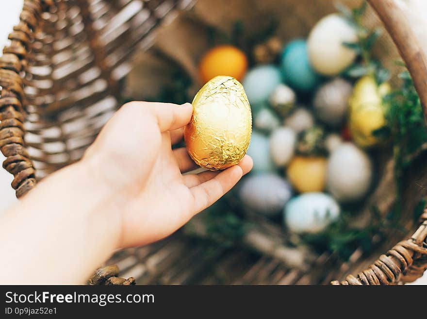 Easter hunt concept. Hand holding golden Easter egg and stylish eggs with green buxus branches in rustic wicker basket on white wooden background. Happy Easter