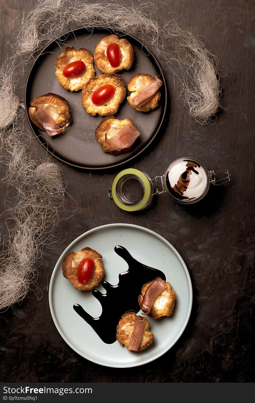 Vertical photo with top view on blue plate with puff pastry cups. Cups are stuffed by melted cheese. Ham and red tomatoes are on baked pastries. Dark sauce is spilled on plate. Vertical photo with top view on blue plate with puff pastry cups. Cups are stuffed by melted cheese. Ham and red tomatoes are on baked pastries. Dark sauce is spilled on plate