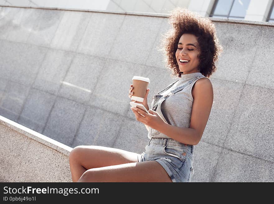 Young woman wearing denim overall shorts in the city street sitting holding cup of hot latte browsing internet on smartphone miling joyful. Young woman wearing denim overall shorts in the city street sitting holding cup of hot latte browsing internet on smartphone miling joyful