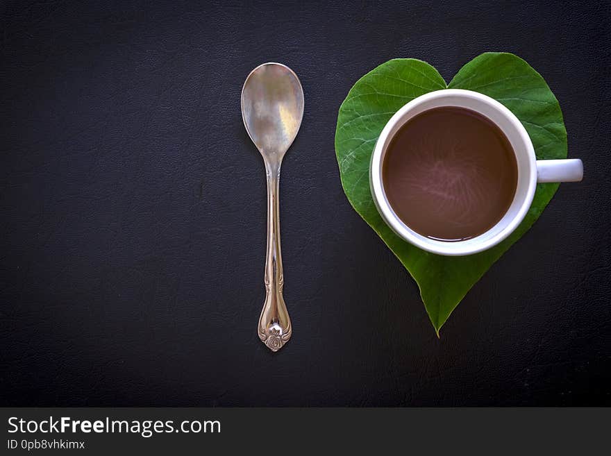 Top view image of a metallic spoon, a heart-shaped green morning glory leaf and a cup of coffee on black leather texture background. Top view image of a metallic spoon, a heart-shaped green morning glory leaf and a cup of coffee on black leather texture background.