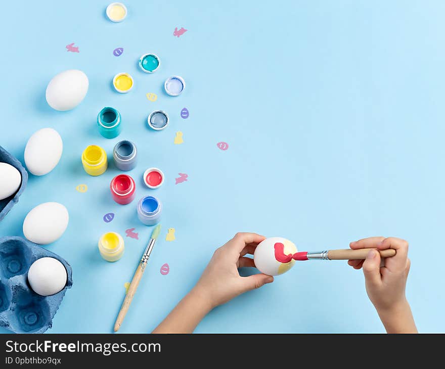 Close up image of kids hands painting an egg. Bottles of various paint, white eggs, bunny and chicken shaped sequins, brushes are around. Top view, space for text. Close up image of kids hands painting an egg. Bottles of various paint, white eggs, bunny and chicken shaped sequins, brushes are around. Top view, space for text.