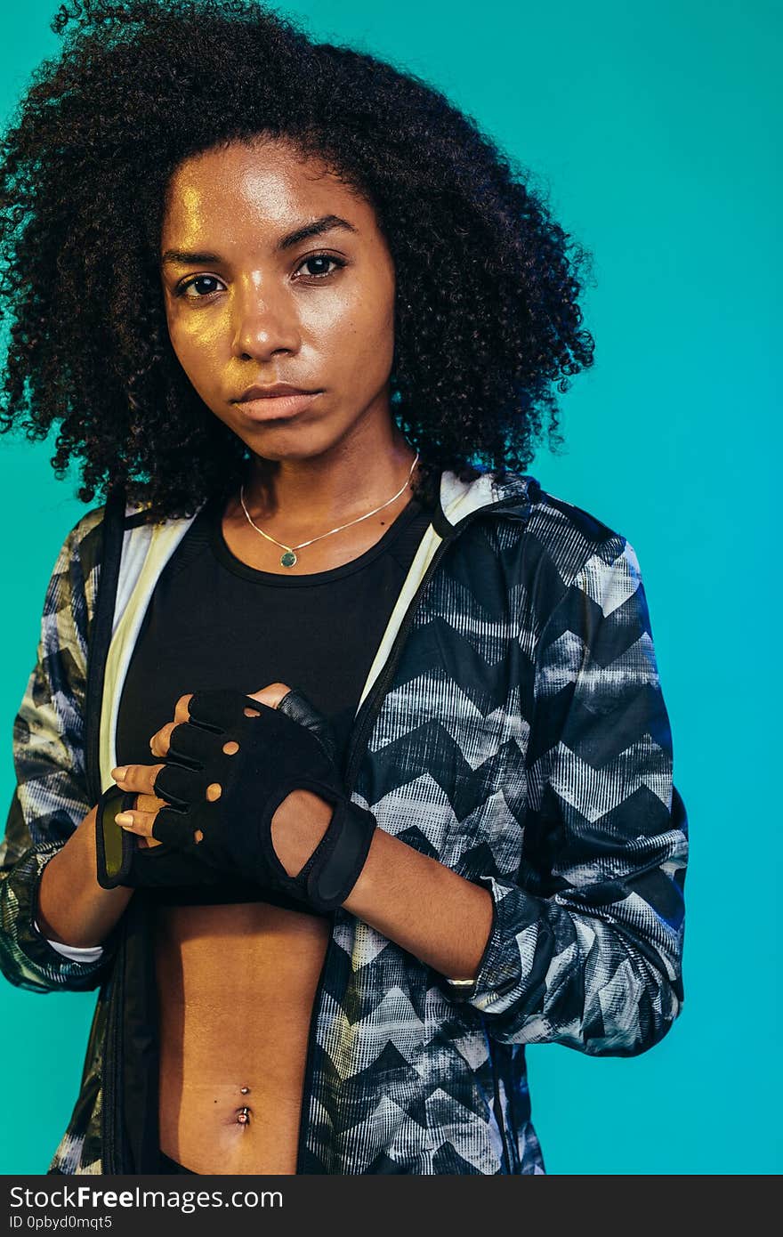 Tough young woman in sportswear standing on blue background. Fit female wearing gloves ready for workout in studio