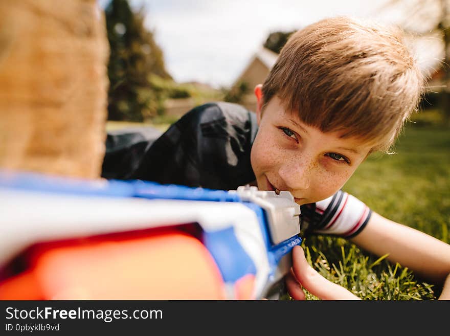 Kid in cape hiding behind a wooden log with toy gun in hands. Cute boy playing with toy gun in playground