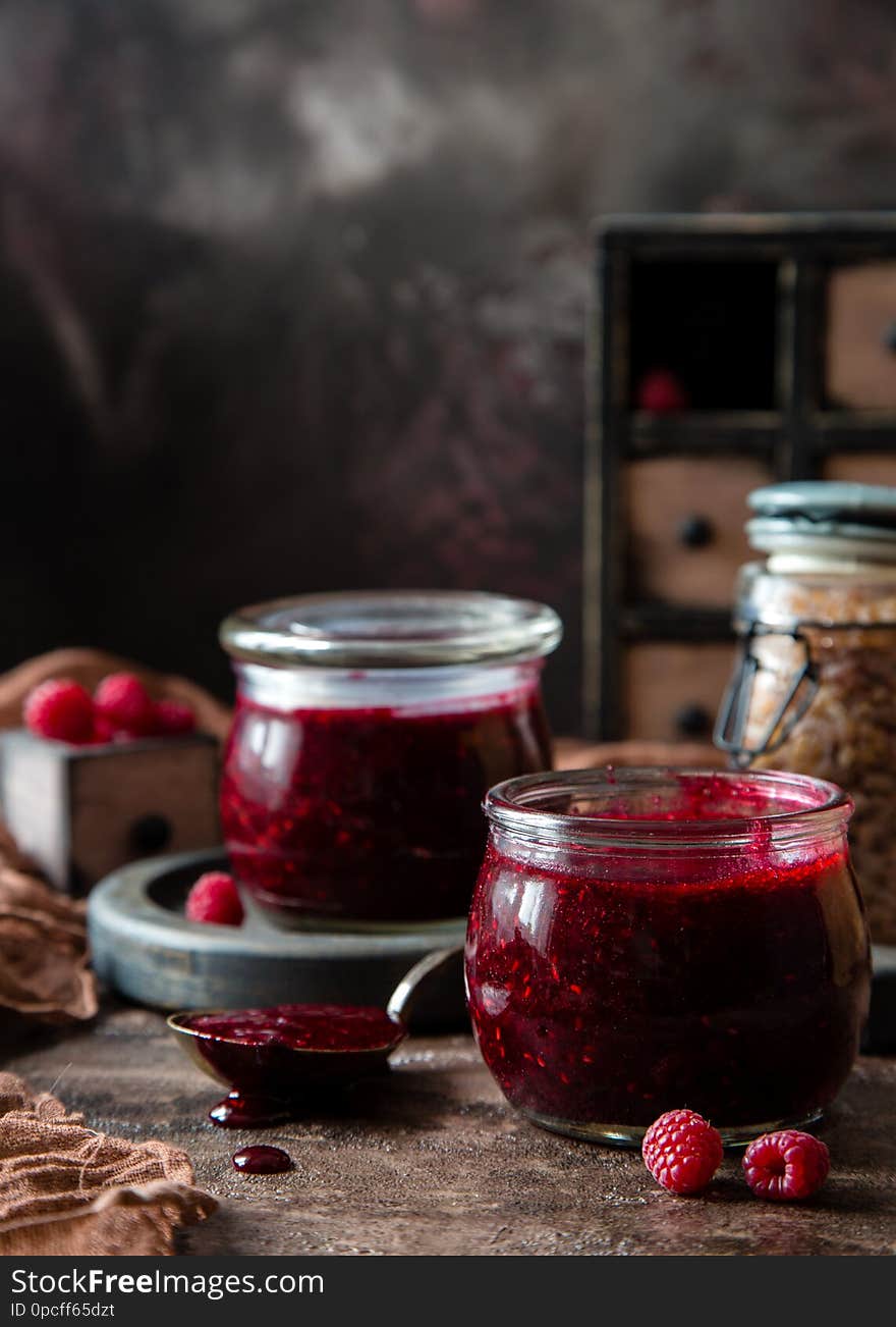 Two glass jars with homemade dark red jam or jelly standing on brown concrete table with raspberries, wooden board, chest of drawers, brown towel and jar with brown sugar. Preservation. Raspberry jam.