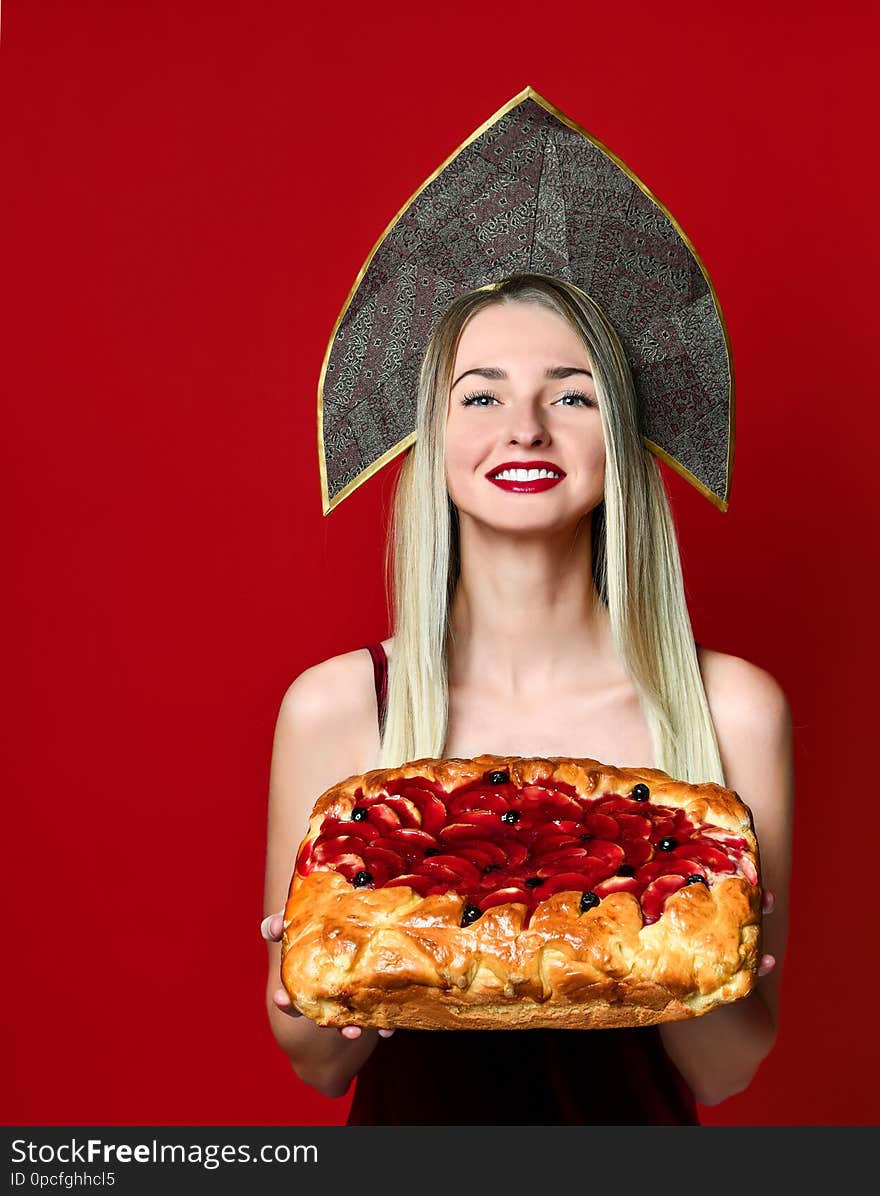 Portrait of a young beautiful blonde in kokoshnik cap holding a delicious homemade berry pie over red background. Portrait of a young beautiful blonde in kokoshnik cap holding a delicious homemade berry pie over red background