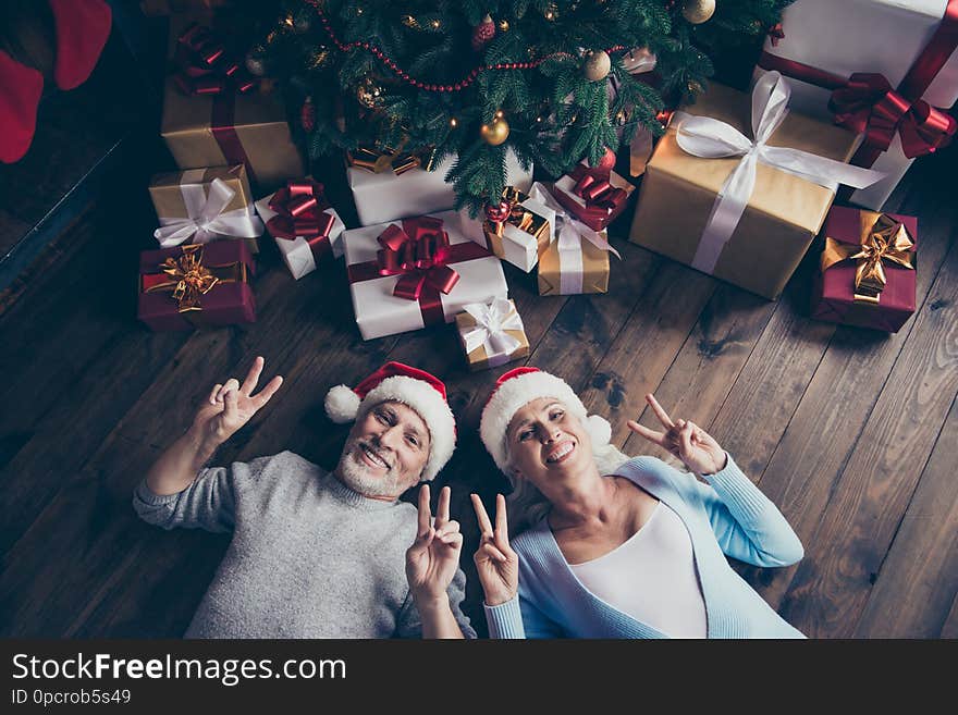 Top above high angle view of two adorable cheerful dreamy funny positive grey-haired spouses lying on floor near decorated fir tree among many bow ribbon boxes showing double v-sign symbol.