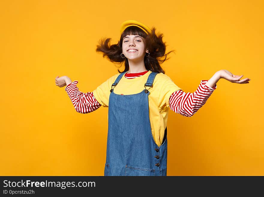 Cheerful girl teenager in french beret denim sundress spreading hands with fluttering hair isolated on yellow wall