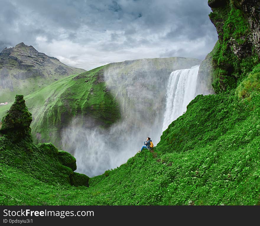 Woman traveler in waterproof clothing is standing on the edge on background of Skogafoss waterfall in Iceland. View from above. Woman traveler in waterproof clothing is standing on the edge on background of Skogafoss waterfall in Iceland. View from above