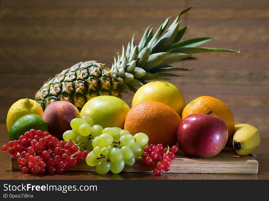 Different kinds of fruits, pineapple, grapes, currant, apple, orange grapefruit, lime, lemon, peach, banana, on the wooden board at the table with brown background. Different kinds of fruits, pineapple, grapes, currant, apple, orange grapefruit, lime, lemon, peach, banana, on the wooden board at the table with brown background.