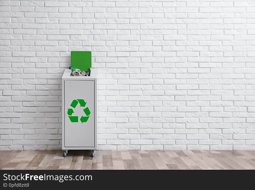 Overfilled trash bin with recycling symbol near brick wall indoors. Space for text