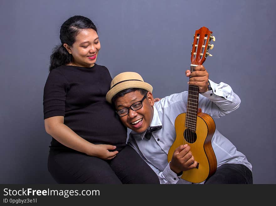 Happy couple expecting baby, men put his ear on wife& x27;s pregnant belly while playing music with an ukulele. Studio shot. Happy couple expecting baby, men put his ear on wife& x27;s pregnant belly while playing music with an ukulele. Studio shot