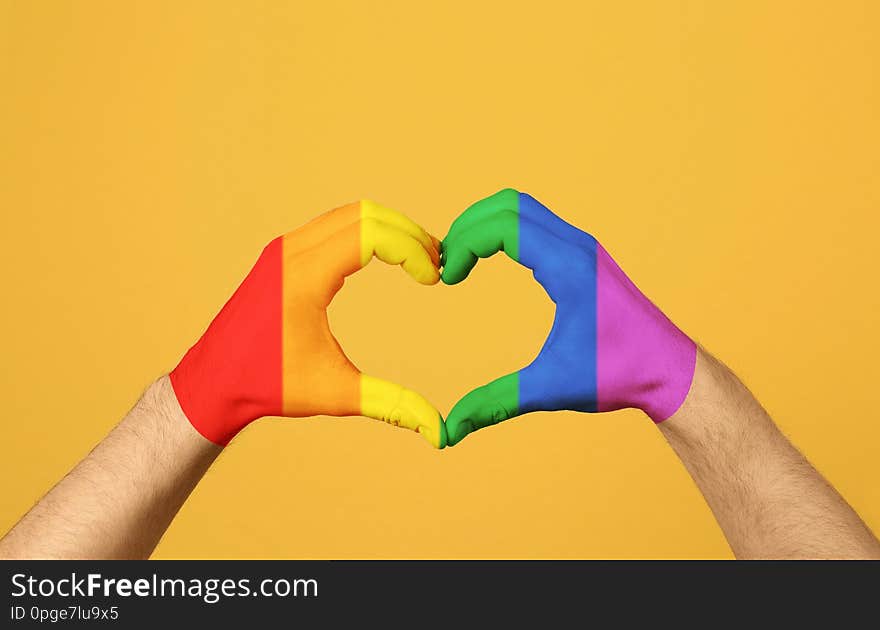 Man making heart with hands painted in LGBT flag on color background, closeup. Gay community