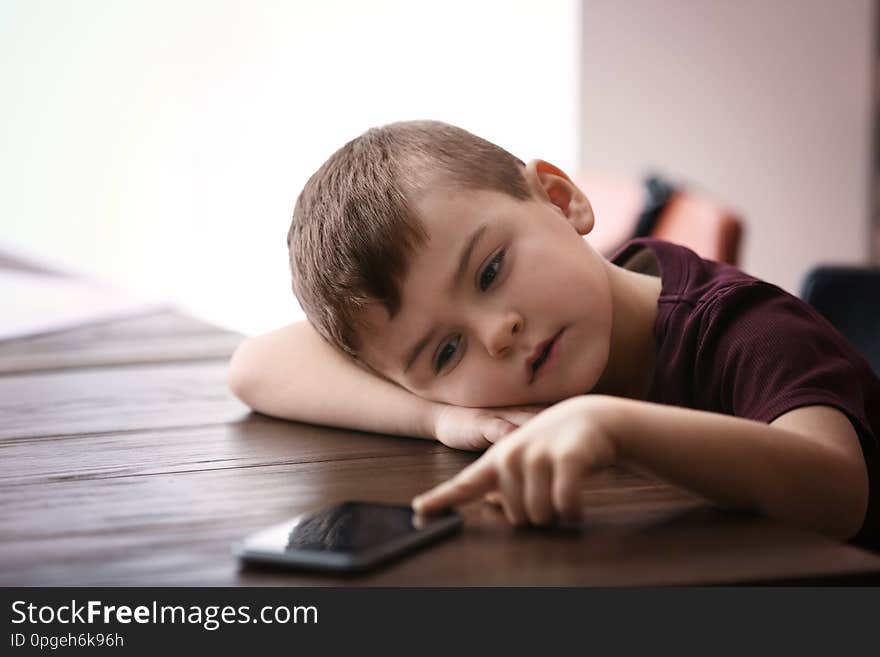 Sad little boy with mobile phone sitting at table indoors