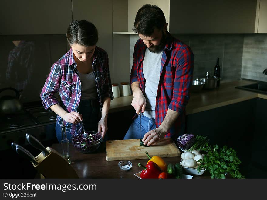 Beautiful young couple in kitchen at home while cooking healthy food. Husband cuts the cucumbers. Wife mix salad. Scene from family life. Horizontally framed shot