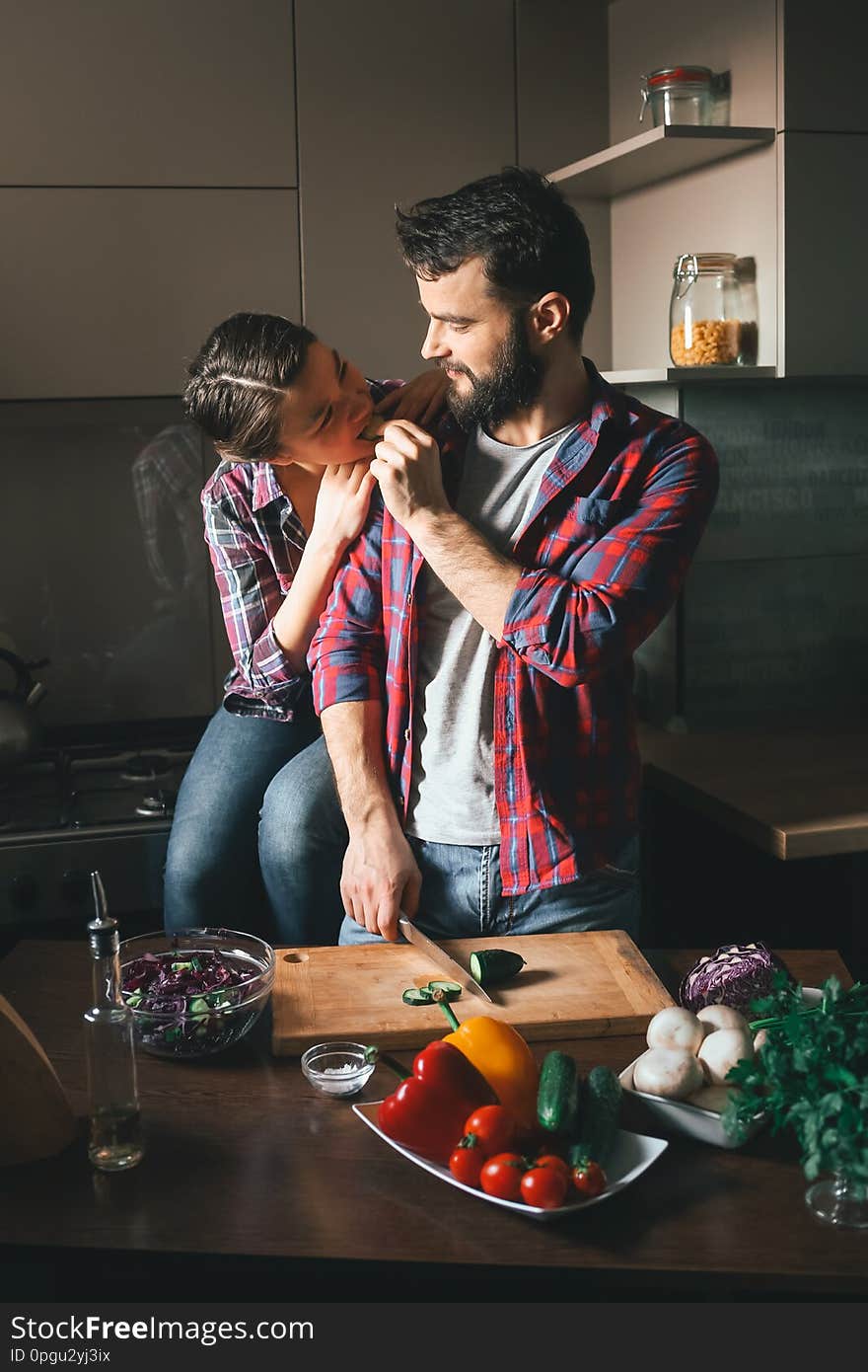 Beautiful young couple in kitchen at home while cooking healthy food. Husband cuts salad and feeds his wife cucumber. Scene from