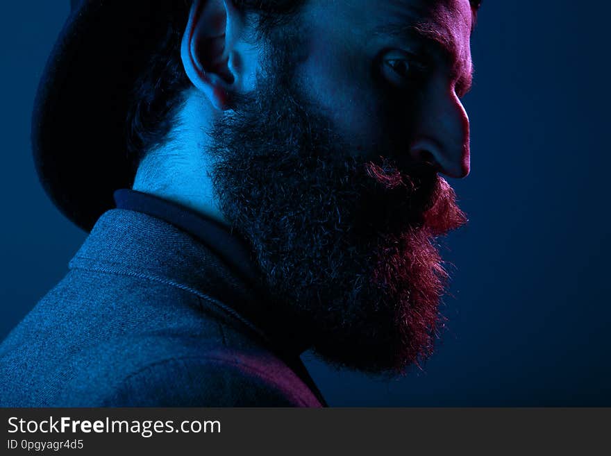 Close up portrait of a bearded man in elegant hat and suit, posing in profile in studio, isolated on blue background.