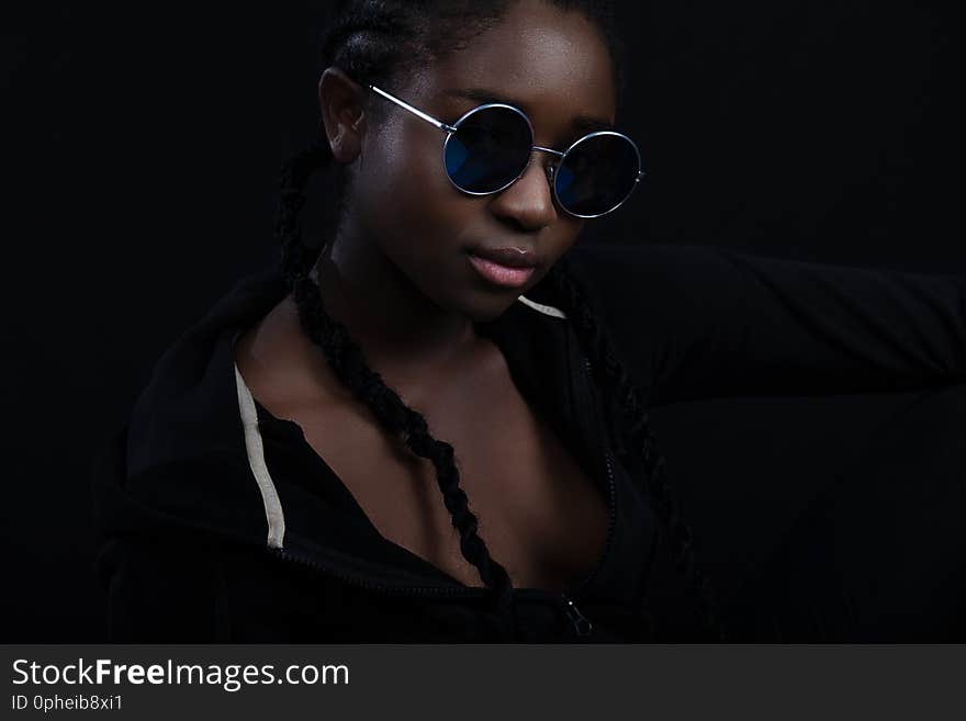 Portrait of a confident and cool young woman with dark skin wearing round sunglasses. High quality low-key studio portrait. Portrait of a confident and cool young woman with dark skin wearing round sunglasses. High quality low-key studio portrait.