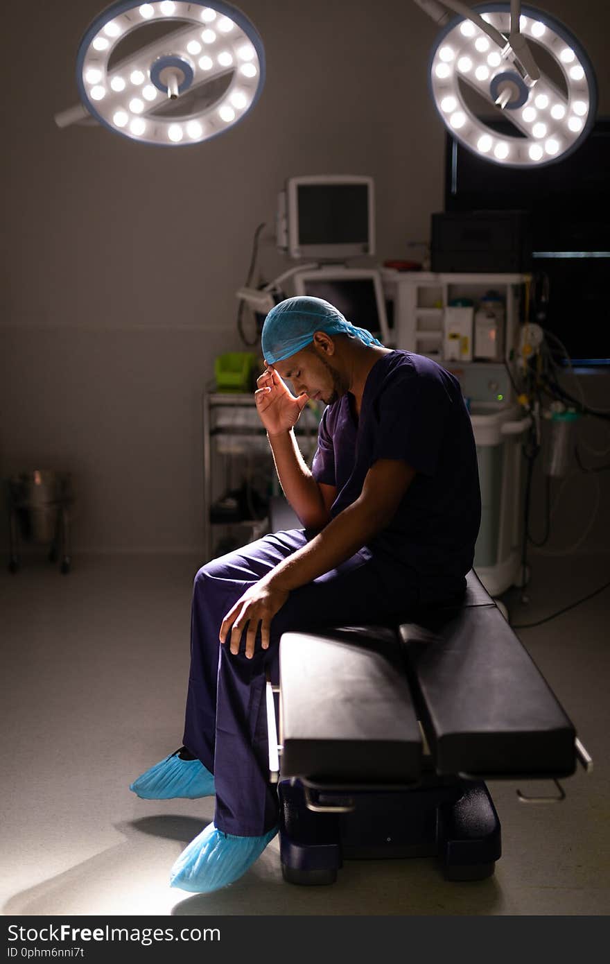 Side view of a nervous African-american male surgeon sitting with his head in his hand in operation room at hospital under a light ray. Side view of a nervous African-american male surgeon sitting with his head in his hand in operation room at hospital under a light ray