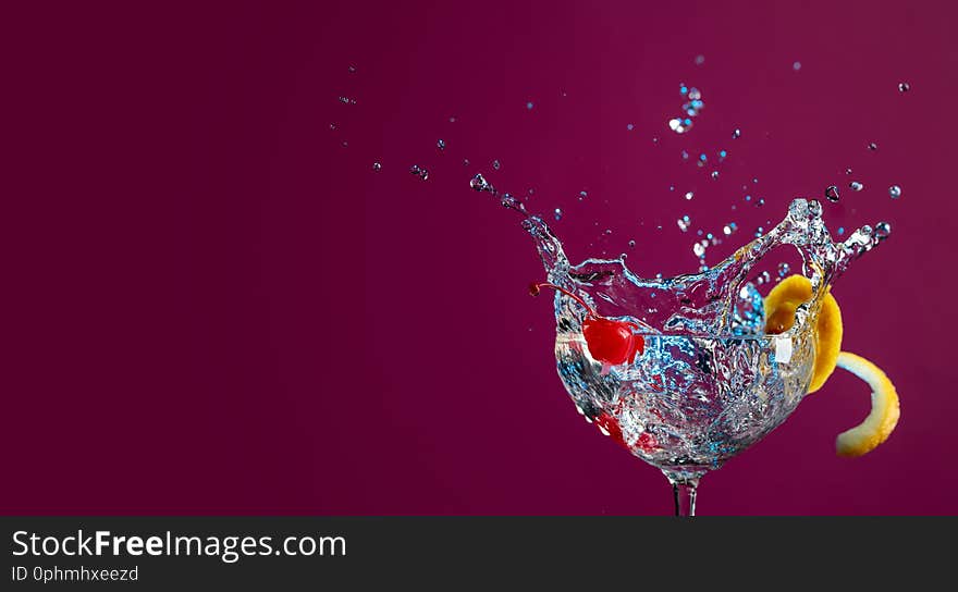 Colorful cocktail in glass with splashes garnished with lemon and cherries. Purple background. Mixed light. Copy space for your text