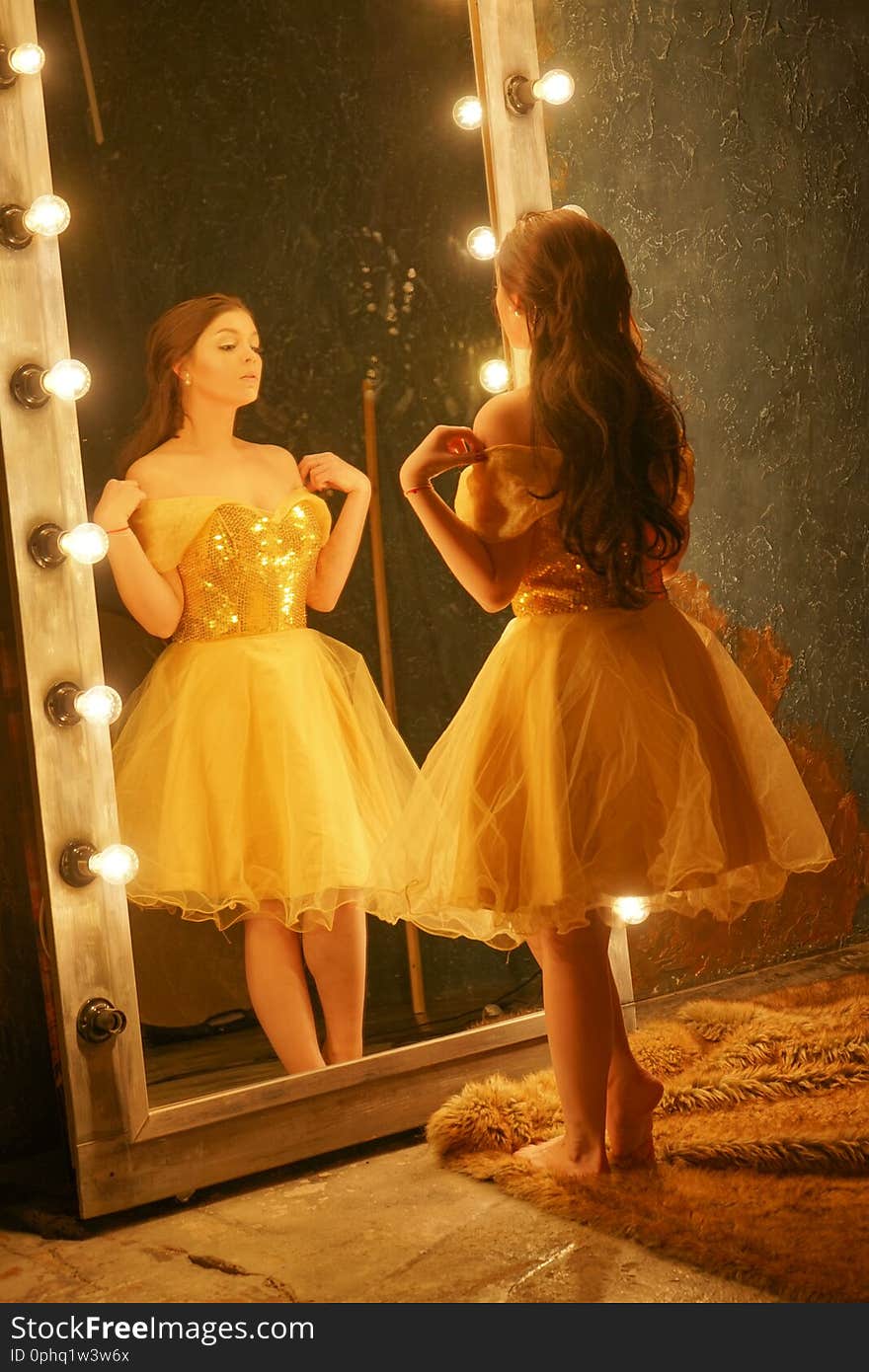 Beautiful young girl in a gold evening dress stands on a fur rug near a large mirror in a frame with lights and looks into her reflection alone