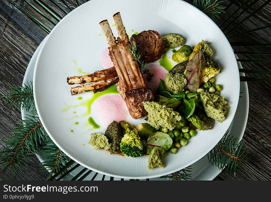 Top view closeup two pieces of grilled ribs with meat served with broccoli, artichokes, green peas and sauce on white restaurant plate on wooden table background. Top view closeup two pieces of grilled ribs with meat served with broccoli, artichokes, green peas and sauce on white restaurant plate on wooden table background