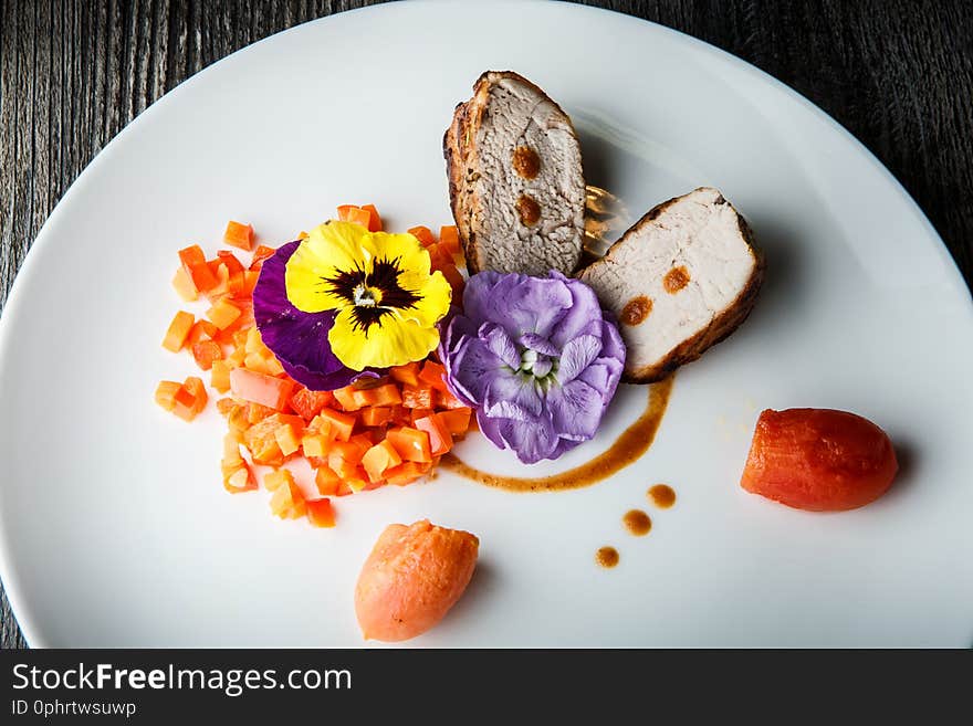 Top view closeup two pieces of baked meat served with sweet pepper and pumpkin salad decorated with flowers on white restaurant plate on wooden table. Top view closeup two pieces of baked meat served with sweet pepper and pumpkin salad decorated with flowers on white restaurant plate on wooden table