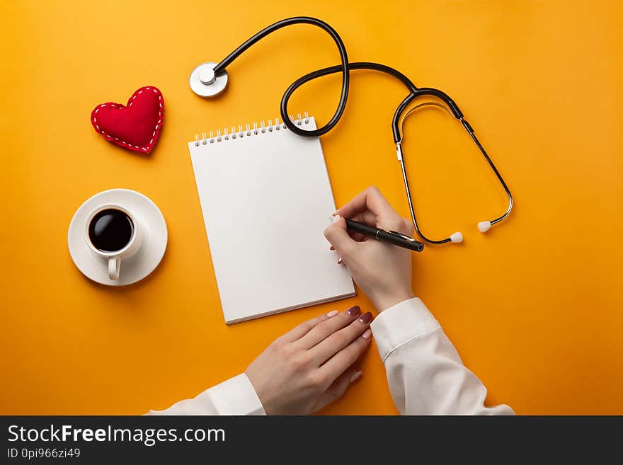 Professional Doctor Writing Medical Records In A Notebook With Stethoscope, Coffee Cup, Syringe And Heart