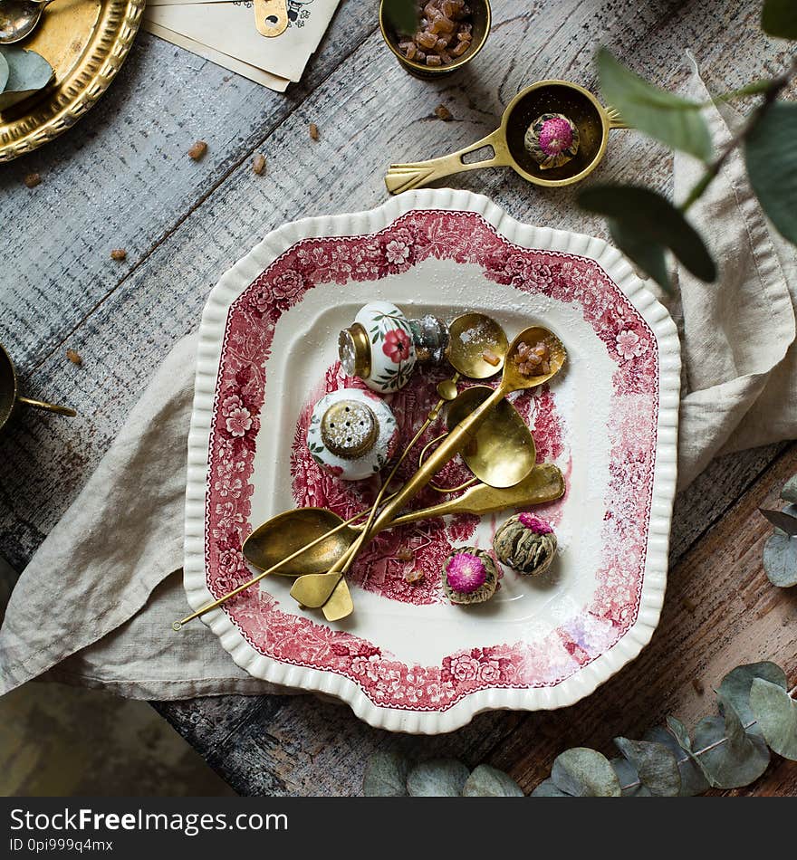 Overhead shot of vintage plate with pink ornament with brass spoons, strainers and chinese tea balls on wooden grey table with brass teapot, cups of brown sugar, branch with leaves and towel. Overhead shot of vintage plate with pink ornament with brass spoons, strainers and chinese tea balls on wooden grey table with brass teapot, cups of brown sugar, branch with leaves and towel