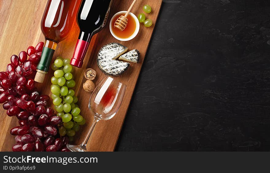 Red and white wine bottles with bunch of grapes, cheese, honey, nuts and wineglass on wooden board and black background. Top view with copy space.