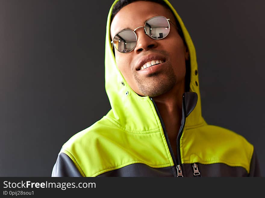 Stylish attractive African American man smiling and looking through mirror sunglasses, wearing green sport hoody, posing on black studio wall. People, emotion and lifestyle concept