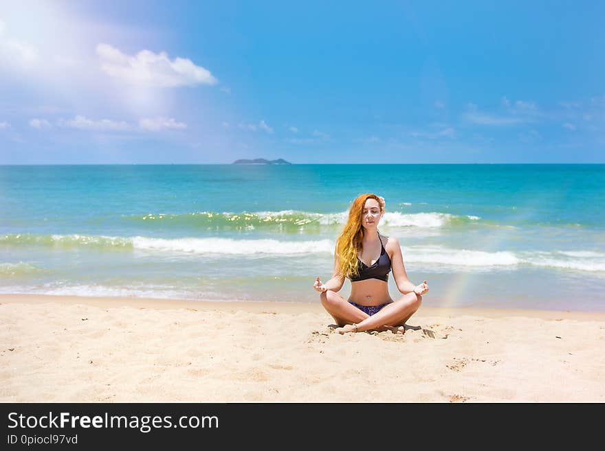 A young, beautiful girl with red hair, in a bikini. practices yoga on the white sand and turquoise sea.