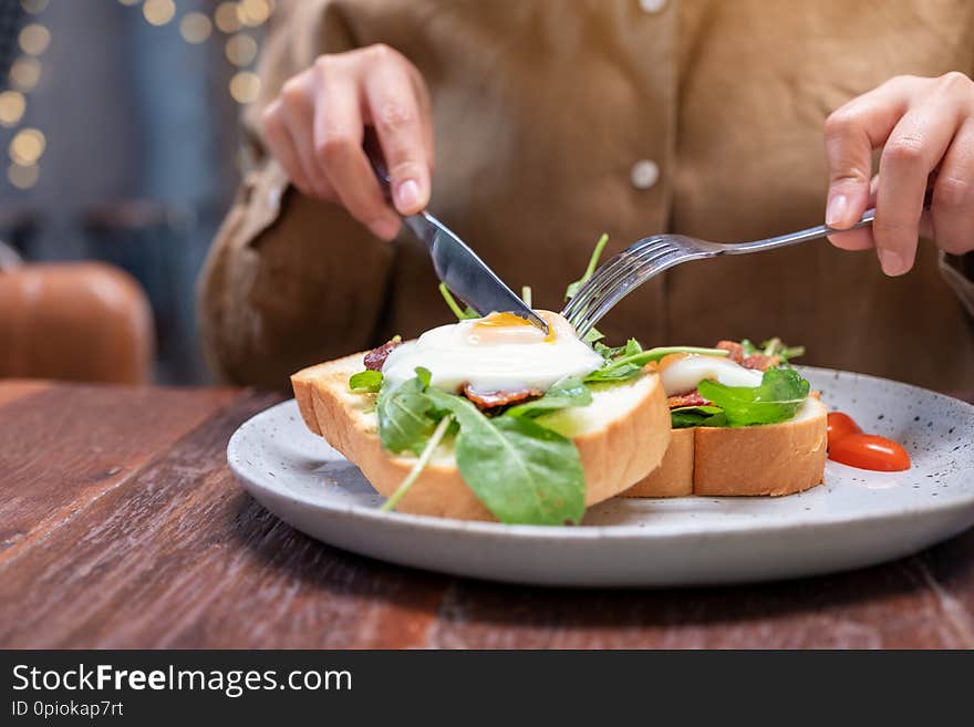 A woman eating breakfast sandwich with eggs, bacon and sour cream by knife and spoon in a plate on wooden table. A woman eating breakfast sandwich with eggs, bacon and sour cream by knife and spoon in a plate on wooden table